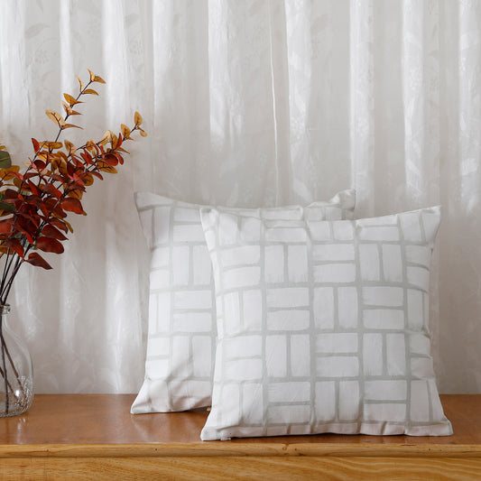 Two white pillows with a checkered pattern, like a picnic blanket. They are sitting on a wooden table next to a vase of flowers.